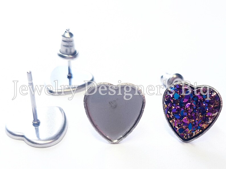 May include: Two heart-shaped earring settings, one with a purple and gold glitter design and the other empty. Both settings are made of silver metal.