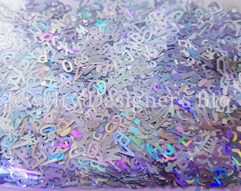 Puede incluir: Una imagen de primer plano de una gran cantidad de purpurina holográfica iridiscente en forma de letras. La purpurina es una mezcla de colores plateado, azul y morado.