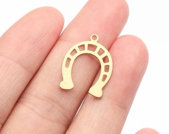 May include: Gold-colored metal horseshoe charm with a cutout design. The horseshoe is held in a hand.