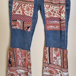 May include: Blue denim jeans with decorative paisley fabric patches on the front pockets and lower legs. The paisley fabric features red, white, and brown colours. The jeans are hanging from a black hanger.