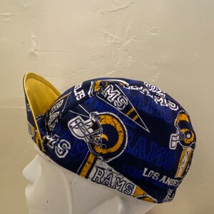 May include: A blue and gold Los Angeles Rams cap. The cap features the team's logo, helmet, and the word "RAMS" in white. The brim is yellow, and the cap is made of a patterned fabric.