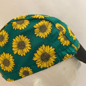 May include: A green cap featuring a repeating pattern of yellow sunflowers. The cap has a black peak and is made of fabric. The sunflowers have detailed yellow petals and dark brown centres. The cap is designed to fit the head.