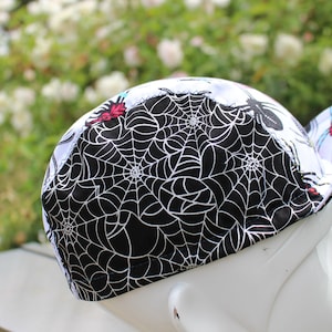 Könnte beinhalten: Eine schwarz-weiße Fahrradmütze mit einem Spinnennetz-Muster und roten und blauen Spinnenillustrationen.