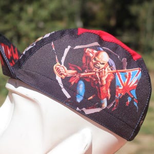 Op de afbeelding: Zwarte en rode pet met een afbeelding van een figuur met een Union Jack-vlag. De pet heeft een gebogen klep en is gemaakt van een duurzame stof. Het ontwerp bevat een rood en zwart patroon op de klep.