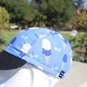 以下が含まれることがあります： アイスクリームコーンとチェリーの柄の青いサイクリングキャップ。キャップは黒いつばと「Hand Made」のラベルが付いています。