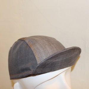 Pode incluir: Uma boné de ciclismo de denim cinza com um detalhe de costura marrom. A boné tem uma aba curva e uma coroa estruturada.