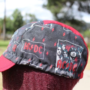 May include: A red and black cycling cap with a black and white AC/DC logo and band member images. The cap has a red brim and a black crown with a repeating lightning bolt pattern.