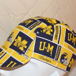 May include: A blue and yellow fabric welding cap with the University of Michigan logo and the words "Wolverine" and "U of M" repeated throughout the design.