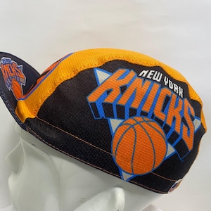 Könnte beinhalten: Eine Radsportmütze mit dem New York Knicks Logo in Blau und Orange auf schwarzem Grund. Die Mütze hat einen gelben Rand an der Oberseite und ein kleines Knicks Logo auf der Unterseite des Schirms. Der Text New York Knicks steht an der Seite.