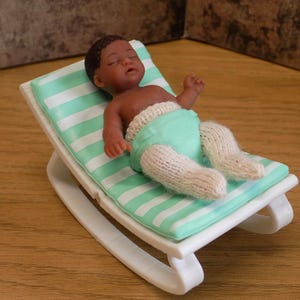 May include: A miniature dark-skinned baby doll with closed eyes, wearing a light green diaper and white knitted leggings, resting on a white plastic rocking chair with a light green and white striped cushion.