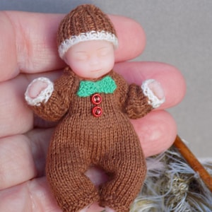 May include: A miniature doll in a brown knitted outfit and hat, with a green bow tie and red buttons. The hat and cuffs have white trim. The doll is about 5 cm tall. A charming, handcrafted item.