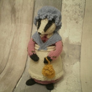 以下が含まれることがあります： 手編みのイタチの置物。青い帽子、ピンクと青のショール、クリーム色のエプロンを着ています。イタチは小さな黄色のハンドバッグを持っています。置物は明るい茶色の表面に立っています。