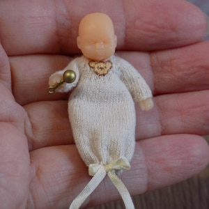 May include: A small, knitted doll baby wearing a white knitted outfit with a gold rattle. The doll has a realistic face and is holding the rattle in its hand.
