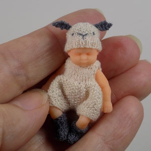 May include: A small, knitted doll dressed in a white, sheep-themed outfit with gray accents. The doll is wearing a hat with ears and a matching onesie.