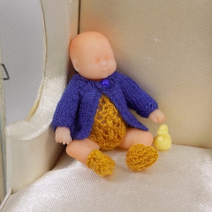 May include: A small knitted doll wearing a purple cardigan, a yellow knitted onesie, and yellow knitted booties. The doll is sitting on a white armchair with a yellow rubber ducky beside it.