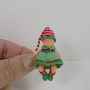 Op de afbeelding: Een kleine gebreide pop met een groen-rode gestreepte muts, een groene jurk met een bruine riem en rood-groen gestreepte sokken.