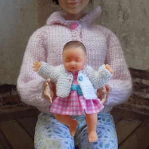 May include: A small doll with closed eyes wearing a white knitted cardigan and a pink and white checkered dress with a blue belt. The doll is held by a larger doll wearing a pale pink knitted sweater and light blue patterned pants.
