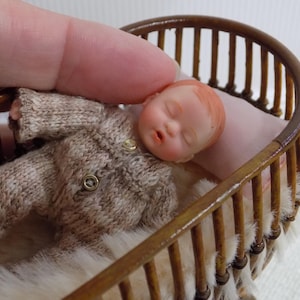May include: A miniature doll with light skin and reddish hair, wearing a beige knitted long-sleeve onesie with two small gold buttons, lying in a dark brown wooden slatted cradle with a soft white faux fur lining and a pale pink pillow.