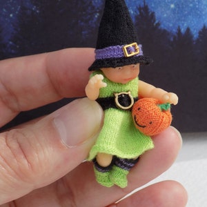 Może przedstawiać: Miniaturowa lalka wiedźmy na Halloween. Nosi czarny kapelusz z fioletową opaską i złotą klamrą, zieloną sukienkę i paski na skarpetkach. Lalka trzyma małą pomarańczową dynię z uśmiechniętą twarzą. Kolekcjonerski przedmiot o tematyce Halloween.