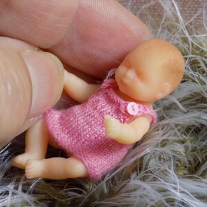 Op de afbeelding: Een kleine, realistische babypop met gesloten ogen, die een roze gebreid jurkje draagt met een knoop. De pop ligt op een pluche, lichtbruine ondergrond.