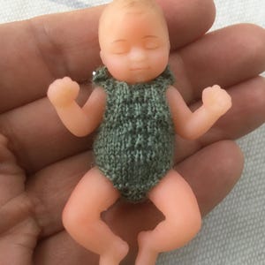 May include: A small baby doll with closed eyes, dressed in a knitted green bodysuit. The doll has a peach skin tone and is posed with arms and legs slightly outstretched. The doll is designed to look like a newborn baby.