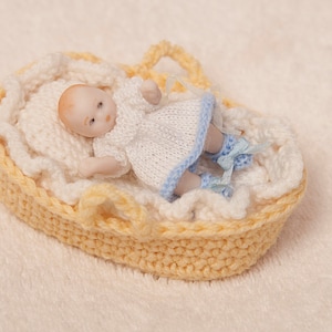 May include: A small porcelain doll wearing a white knitted dress with blue trim, lying in a yellow crocheted basket.