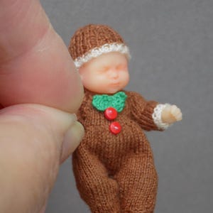 May include: A miniature doll dressed in a brown knitted outfit with a matching hat. The doll has a white trim on the hat and sleeves, a green collar, and two red buttons. The doll is held by a person's fingers.