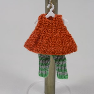 May include: A miniature knitted outfit, featuring an orange top and striped green and gray pants, hangs on a white hanger. The outfit is displayed on a wooden stand against a white background. The knitted texture is visible.