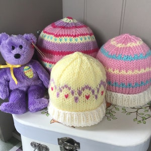 May include: Three knitted hats in pastel colors. The hats are yellow, pink, and purple with white stripes. The hats are sitting on a white surface with a teddy bear in the background.