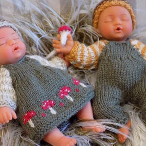 Puede incluir: Dos muñecas bebé vestidas con trajes de punto hechos a mano. Una muñeca lleva un vestido gris con diseños de setas rojas y blancas, un sombrero marrón y mangas blancas. La otra muñeca lleva un peto gris y un jersey a rayas naranja y blanco, con un gorro a juego.