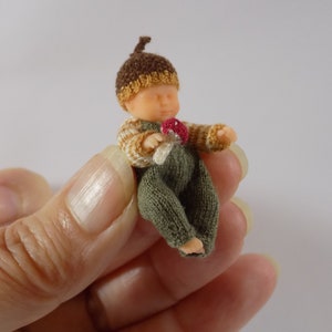 May include: A small knitted doll wearing a brown and yellow acorn hat and green overalls. The doll is holding a small red and white mushroom.