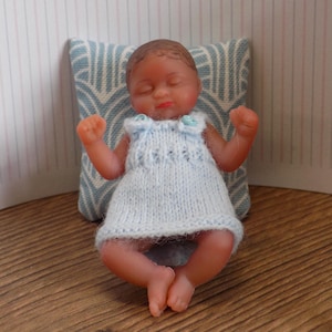May include: A miniature baby doll with brown skin, wearing a light blue knitted dress. The doll is resting against a patterned pillow. The baby doll has its eyes closed and arms raised.