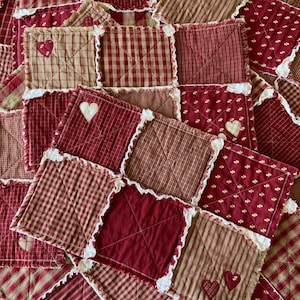 May include: A patchwork quilt featuring squares in various shades of red and tan, with white heart appliqués and decorative stitching. The quilt has a homespun, country aesthetic, with a mix of plaid, striped, and solid fabrics.