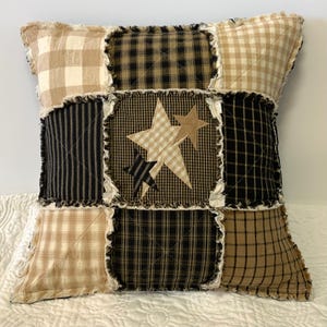 May include: Square patchwork pillow with a star design. The pillow features a mix of tan, black, and cream fabrics in plaid, striped, and solid patterns. The center square has tan and black stars.