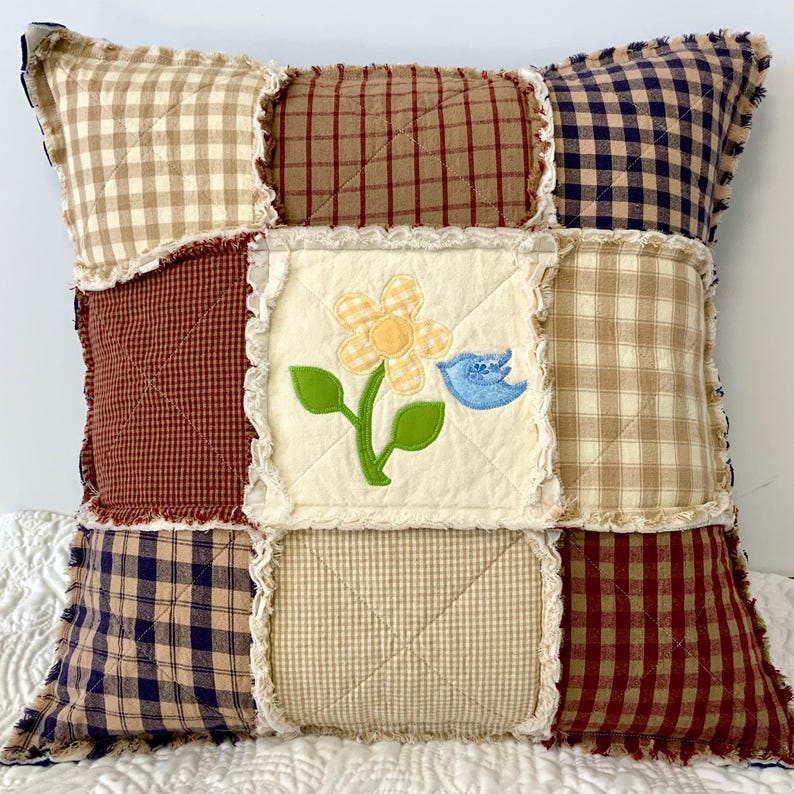 Peut inclure: Un coussin patchwork avec un motif floral et un oiseau au centre. Le coussin est fait de tissus &agrave; carreaux de diff&eacute;rentes couleurs dans les tons de brun, bleu et beige.