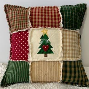 May include: A patchwork pillow with a Christmas tree design in the center. The pillow is made of various fabrics in red, green, and beige with a white background. The tree is green with a red heart and a yellow star on top.