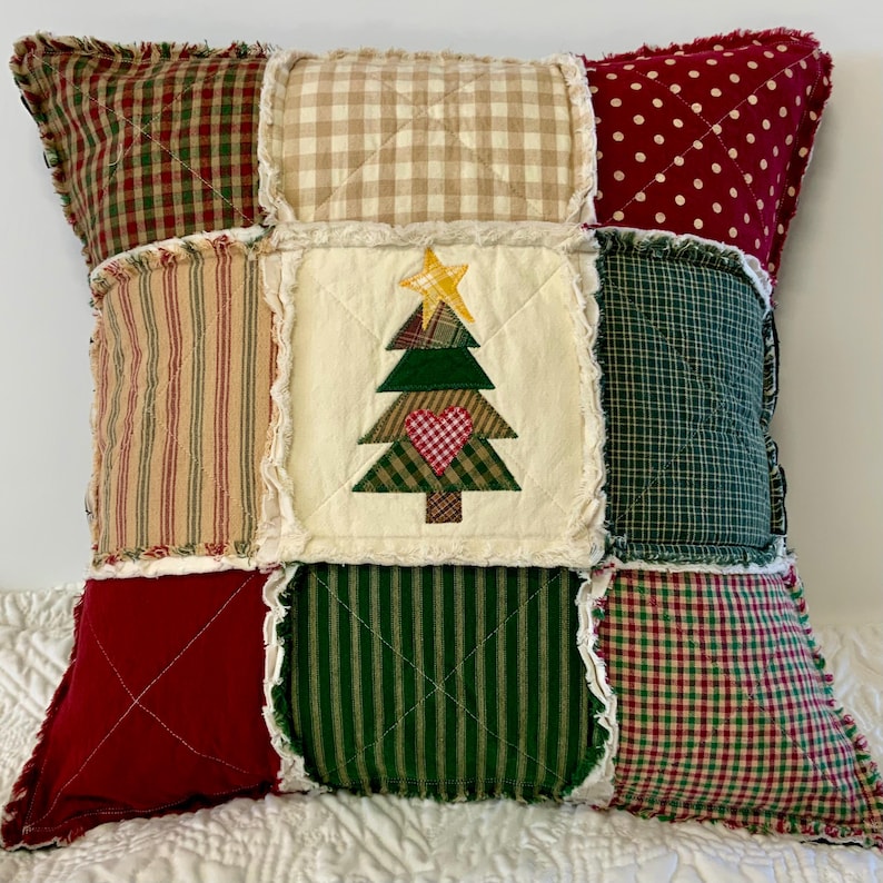 May include: A patchwork pillow with a Christmas tree design in the center. The pillow is made of various fabrics in red, green, white, and brown. The tree is green with a red heart and a yellow star.