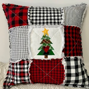 May include: A patchwork pillow with a Christmas tree design in the center. The pillow is made of red and black plaid fabric, white and black gingham fabric, and white fabric. The tree is green with a brown trunk and a yellow star on top. There is a red heart on the tree.