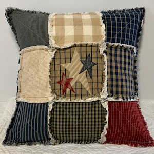 May include: Square patchwork pillow with a variety of fabrics in navy, tan, and red. The center features a tan square with three star appliqués. The pillow has a fringed edge and a quilted design. A decorative accent for a bed or sofa.