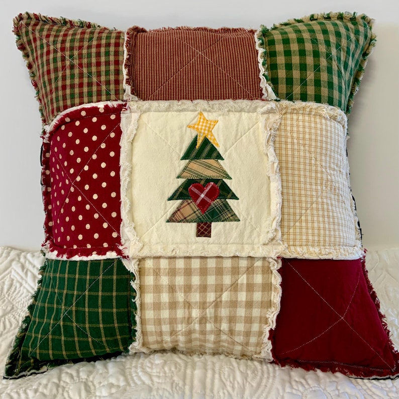 May include: A patchwork pillow with a Christmas tree design in the center. The pillow is made of red, green, white, and brown fabrics with a frayed edge.