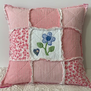 May include: A pink and white patchwork pillow with a blue flower and a heart applique. The pillow has a frayed edge and is made of different fabrics.