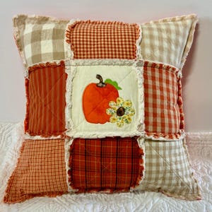 May include: A square rag quilt pillow featuring a patchwork design of orange, red, and beige plaid and gingham fabrics. The center square is cream-colored and displays an orange pumpkin appliqué with a small yellow and white floral accent.