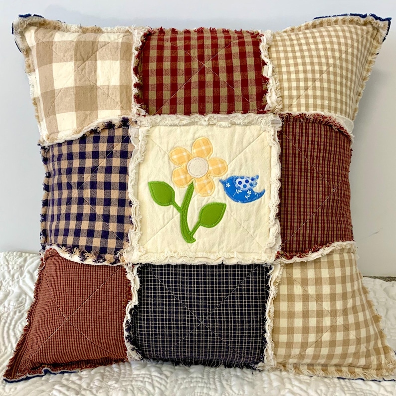 Peut inclure: Un coussin patchwork avec une applique florale et un oiseau. Le coussin est fait de diff&eacute;rents tissus vichy de couleur rouge, bleu et beige.