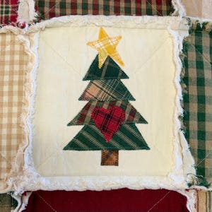 May include: A white fabric square with a patchwork Christmas tree design. The tree is made of green, brown, and red plaid fabric and has a yellow star on top. A red heart is in the center of the tree.