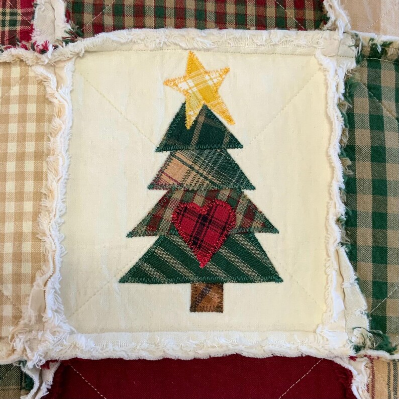 May include: A white fabric square with a patchwork Christmas tree design. The tree is made of green, brown, and red plaid fabric and has a yellow star on top. A red heart is in the center of the tree.