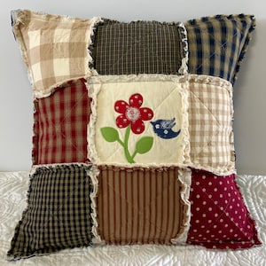 May include: A square rag quilt pillow featuring a nine-patch design with frayed seams. The fabric squares include beige and white gingham, navy and black check, red and white plaid, red and white polka dots, and a central cream square with a red flower and blue bird applique.
