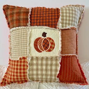 May include: Square decorative pillow with a patchwork design. The pillow features a central square with an embroidered pumpkin. The surrounding squares have various plaid and striped patterns in shades of orange, brown, and cream. The edges have a frayed, textured trim.