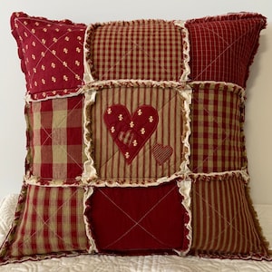 May include: A red and brown patchwork pillow with a heart design. The pillow is made of different fabrics, including plaid and striped fabrics. The heart is embroidered with a small heart inside.