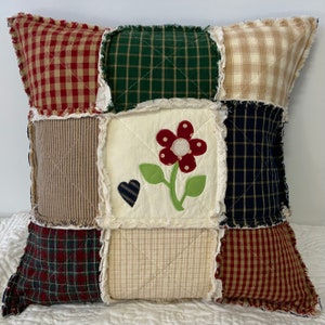 May include: A patchwork pillow with a red flower applique and a heart. The pillow is made of various plaid fabrics in shades of red, green, blue, and beige.
