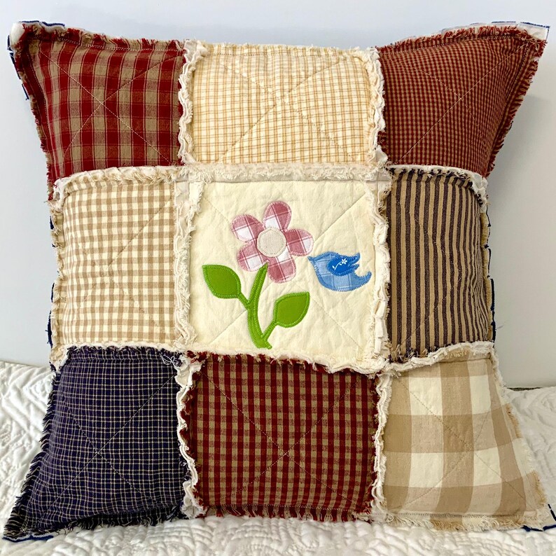 Peut inclure: Un coussin patchwork avec un motif floral et un oiseau bleu. Le coussin est fait de diff&eacute;rents tissus dans des tons de brun, beige, rouge et bleu. Le coussin a un bord effiloch&eacute;.